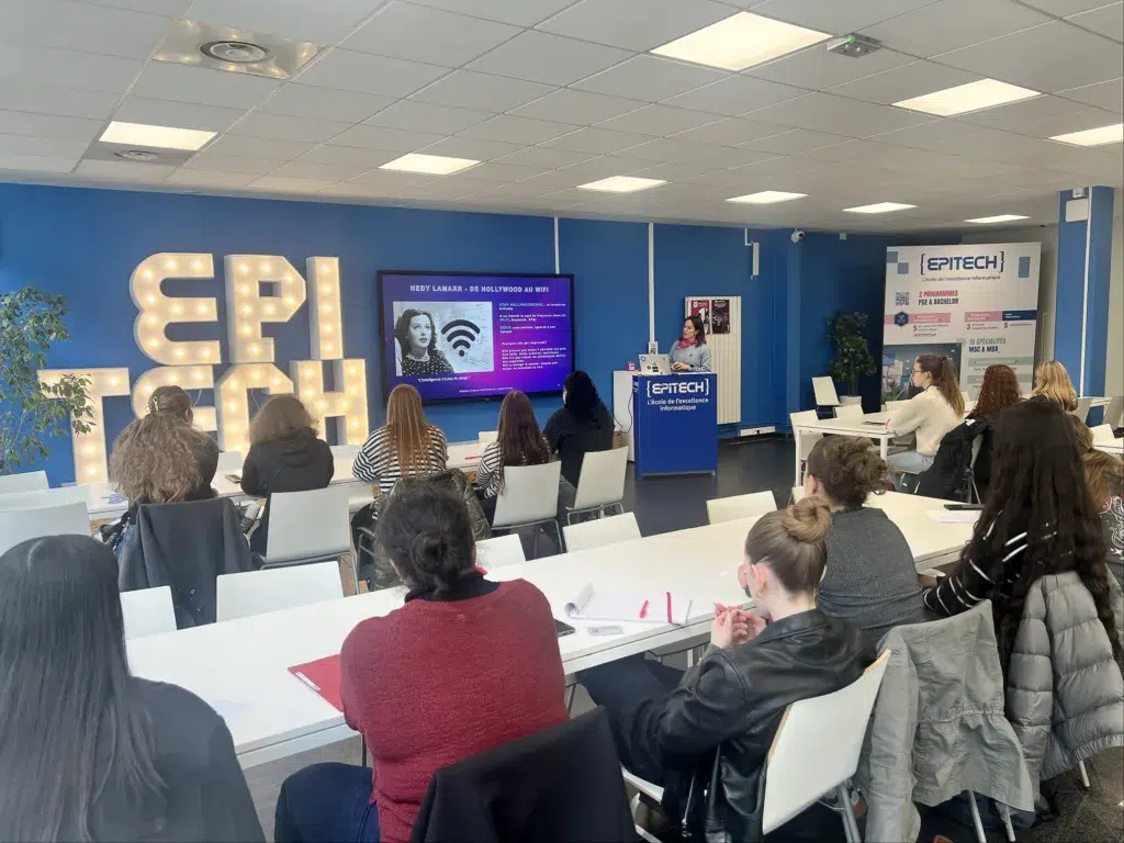 Présentation à l'école EPITECH avec des étudiants assis dans une salle de cours, écoutant une intervenante près d'un pupitre et d'un grand écran.