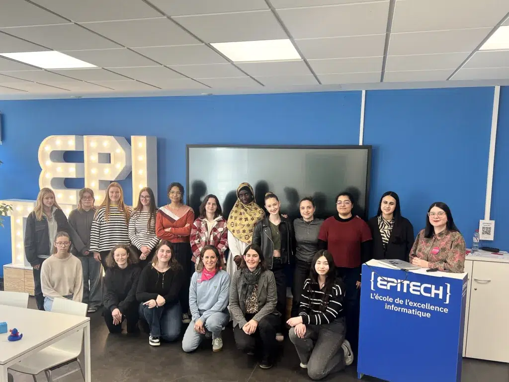Groupe diversifié de jeunes femmes, étudiantes à EPITECH, posant pour une photo de groupe dans un espace d'apprentissage moderne, avec le logo de l'école en arrière-plan.